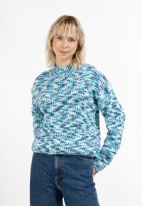 Kék árnyalatokban készült kötött pulóver, magas nyakkivágással, hosszú ujjakkal és texturált cikk-cakk mintával. Magas derekú farmernadrággal párosítva.