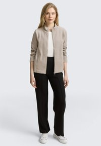 Giacca chiara beige con zip e finitura testurizzata, maniche lunghe e due tasche laterali, abbinata a pantaloni neri su misura e sneakers bianche.