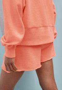 Koraalkleurige sweatshirt en shorts set, gemaakt van zacht materiaal. De sweatshirt heeft een geribbelde onderkant en manchetten; de shorts hebben een golvende zoom.