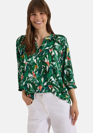 Mujer sonriendo, lleva una blusa verde con estampado de hojas tropicales y flores, y pantalones blancos, de pie con una mano en el bolsillo.