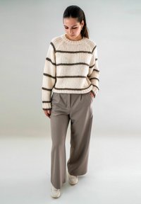 Warm crème trui met bruine strepen, geribbelde textuur. Lichtgrijze brede broek van gladde stof, en witte sneakers maken de outfit compleet.