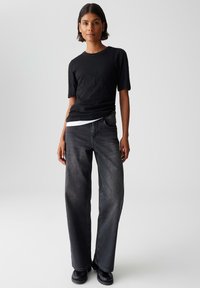 Donna in piedi con una maglietta nera a maniche corte, una maglietta bianca sotto, jeans larghi neri sbiaditi e scarpe nere su uno sfondo neutro.