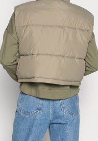 Beige puffervest med en glat finish, der har vandrette syninger, en høj krave og en elastisk kant, båret over en grøn sweatshirt.