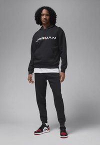 Mustapaita, jossa on "JORDAN"-logo, kengurupocket ja nauhahiha; yhdistettynä samanvärisiin mustiin verkkareihin ja musta/punaisiin lenkkareihin.