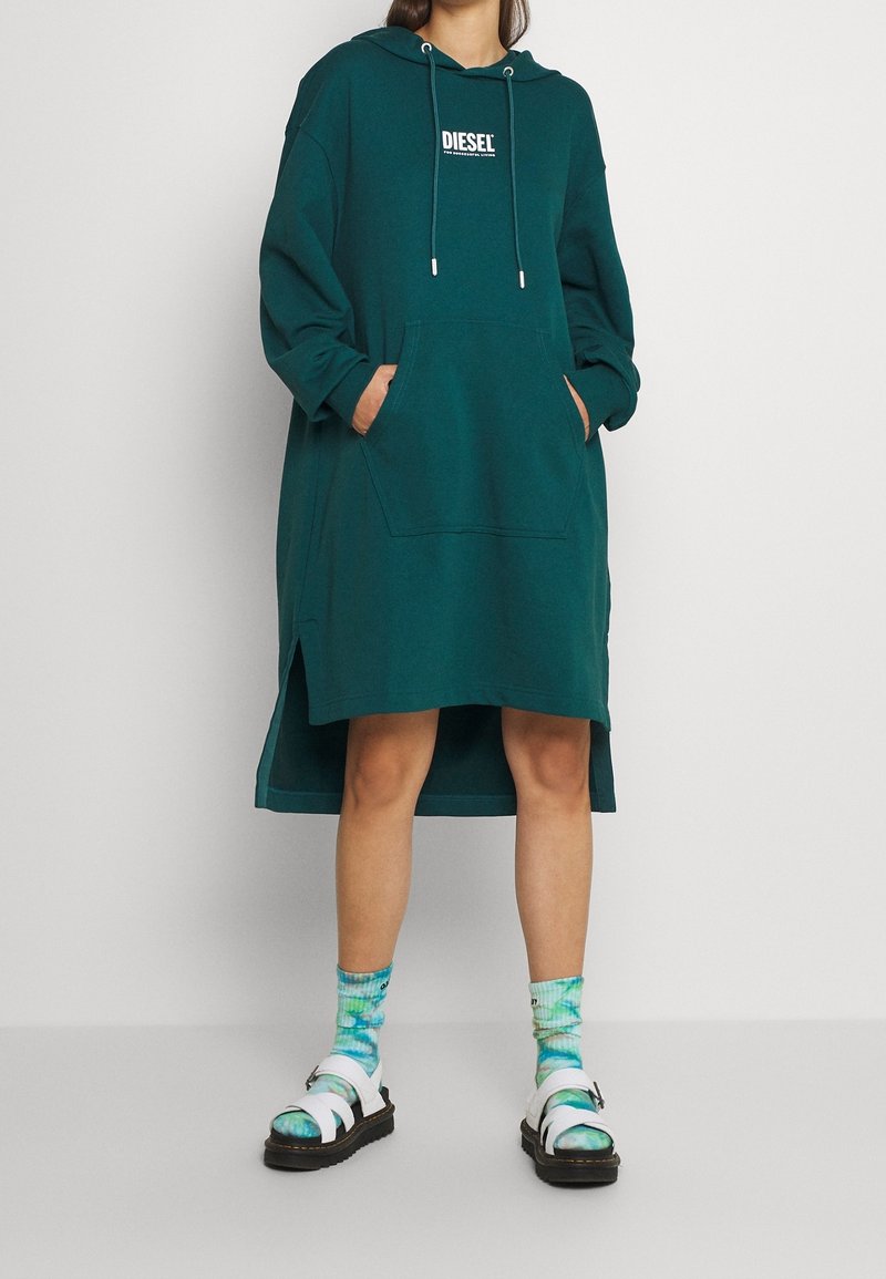 Robe à capuche surdimensionnée couleur sarcelle avec poche frontale, fentes sur les côtés et détail logo blanc ; associée à des chaussettes tie-dye et des sandales chunky.