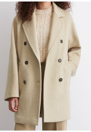Vrouw draagt een beige, dubbelgeknoopte wollen jas over een grove gebreide trui, met één hand in de jaszak, tegen een effen achtergrond.