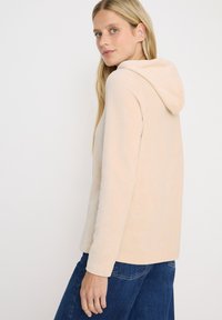Femme aux longs cheveux blonds portant une veste à capuche beige clair et un jean bleu, de profil avec une expression neutre.