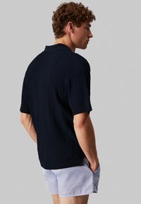 Mörkblå kortärmad polo med en texturerad stickad mönster och en ribbad krage, bärs med ljusgrå shorts.