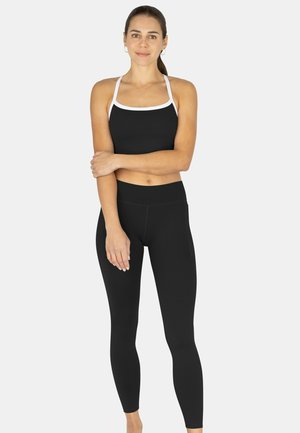 Junge Frau steht, trägt schwarze Leggings und ein schwarzes Crop-Top mit weißen Trägern, Arme verschränkt und leicht lächelnd.