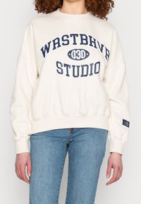 Persona vestida con un sudadera color crema que dice "WRSTBHVR 03D STUDIO" en letras azul marino y unos jeans celestes, de pie con los brazos relajados.