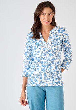 Frau trägt eine weiße Bluse mit langen Ärmeln und blauem Blumenmuster sowie hellblaue Jeans, steht vor einem schlichten hellen Hintergrund.