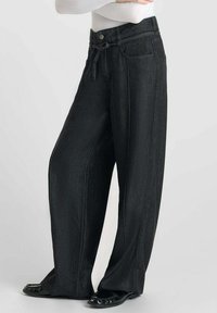Zwarte high-waisted denim broek met wijde pijpen, een trekkoord in de taille, voorzakken, verticale naden en een gladde textuur. Zwarte schoenen worden eronder gedragen.
