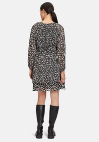 Schwarz-weiß gemustertes Kleid mit langen, durchsichtigen Ärmeln, tailliertem Bund und gestuftem Saum. Getragen mit kniehohen schwarzen Lederstiefeln.