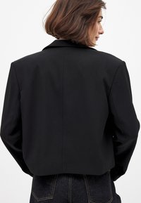 Moteris su trumpais rudais plaukais, vilkinti juodą trumpą švarką ir tamsias džinsines kelnes, matoma iš nugaros prieš paprastą baltą foną.