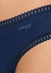 Navy blue Sloggi briefs made of smooth fabric. Features a contrasting lace trim at the waistband with scalloped edges. Subtle logo printed.