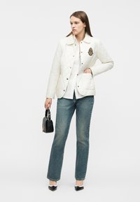 Γυναίκα που φοράει λευκό καπιτονέ μπουφάν με έμβλημα, λευκό πουκάμισο, μπλε τζιν, μαύρα μυτερά παπούτσια, κρατώντας μια μικρή μαύρη τσάντα χειρός με ιμάντα.