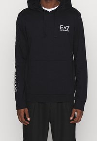 Sudadera con capucha negra hecha de tejido suave, que cuenta con un bolsillo frontal, cordones y una marca en el pecho y la manga.