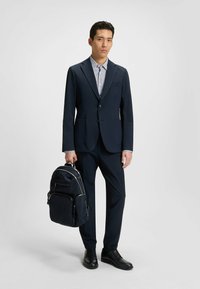 Marinblå kostym med en figurnära jacka och matchande byxor, tillsammans med en mönstrad skjorta. Bär en mörk ryggsäck; har på sig svarta skor.