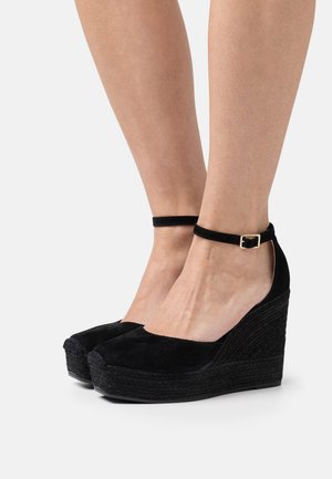 Femme portant des talons compensés en suède noir avec des brides autour de la cheville et des boucles dorées, montrée de mi-mollet vers le bas sur un fond blanc.