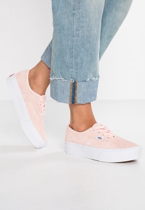 Voeten met lichtroze suède Vans sneakers en cropped lichtblauwe spijkerbroeken met versleten zomen op een witte vloer.