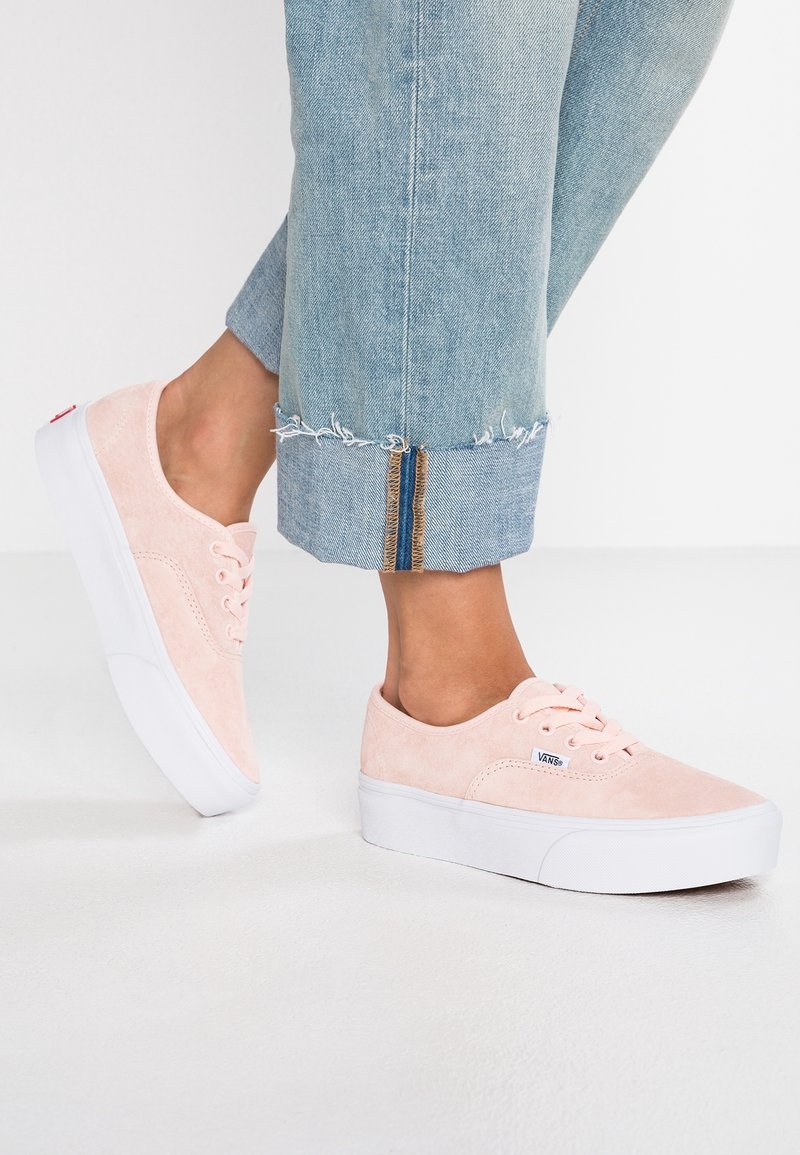 Pieds portant des baskets Vans en daim rose clair et un jean denim bleu clair raccourci avec des ourlets effilochés sur un sol blanc.