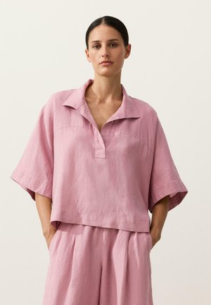 Femme aux cheveux foncés attachés, portant un haut ample à manches courtes rose pâle et un pantalon large assorti, debout les mains dans les poches.