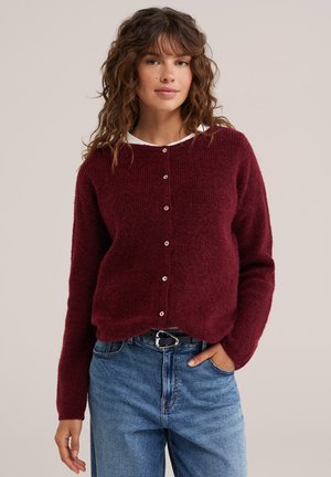Junge Frau mit lockigem Haar, die einen burgunderfarbenen, geknöpften Pullover über einem weißen Hemd und blauen Jeans trägt, steht mit einer Hand in der Hosentasche.