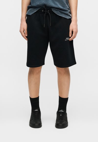 Personne portant un short noir à cordon avec un petit texte blanc sur la jambe gauche, des chaussettes noires et des baskets noires sur un fond uni.