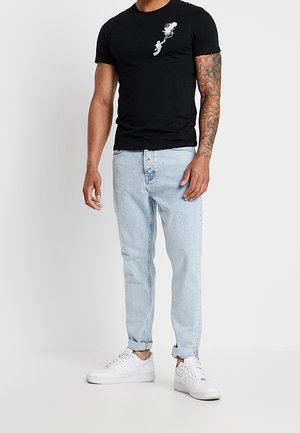 Man draagt zwart T-shirt met kleine Mickey Mouse afbeelding op de borst, lichtblauwe opgerolde spijkerbroek, witte sneakers en een getatoeëerde rechterarm.