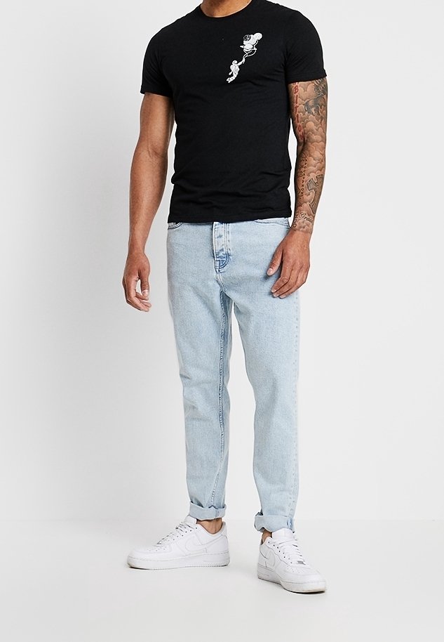 Homme portant un T-shirt noir avec un petit motif de Mickey Mouse sur la poitrine, un jean bleu clair retroussé, des baskets blanches et un bras droit tatoué.