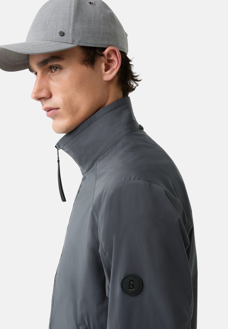 Graue Nylonjacke mit hohem Kragen und Reißverschluss, ausgestattet mit einem runden schwarzen Logoabzeichen am Ärmel. Ergänzt durch eine graue Mütze.