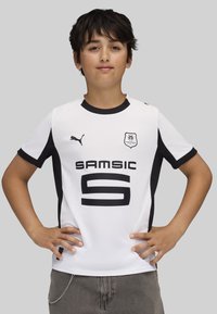 Camiseta deportiva blanca con acentos en negro; cuenta con cuello redondo, mangas cortas y un bold texto negro que dice "SAMSIC 5". Logotipo de la marca en el lado izquierdo del pecho.