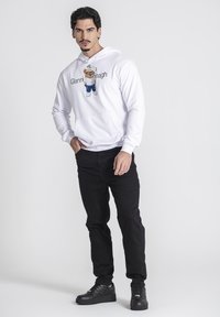 Sudadera blanca con un gráfico de oso bordado, jeans negros y zapatillas negras. Presenta un bolsillo frontal y capucha con cordón. Diseño simple.