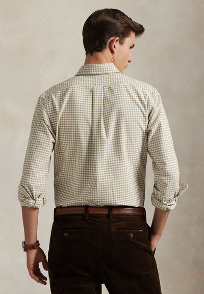 Bărbat cu mânecile suflecate, purtând o cămașă în carouri beige și alb, băgată într-o pereche de pantaloni maro închis, cu o curea maro din piele, cu fața spre spate.