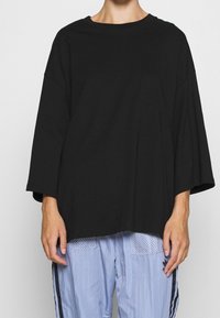 Eine Person trägt ein übergroßes schwarzes Langarmshirt, kombiniert mit hellblauen Adidas-Trackpants mit schwarzen Streifen und Netzseitentaschen.