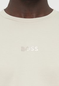 Svetlo bež bombažna majica s tkanim rebrastim okroglim ovratnikom; na sprednji strani ima subtilno vdelan teksturiran logotip "BOSS".