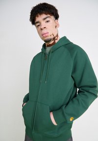 Grüne Reißverschlussjacke mit Kapuze und Kordelzug, Fronttaschen und Kontrastnähten. Verfügt über ein Logo-Patch am Ärmel. Weicher, dicker Stoff.
