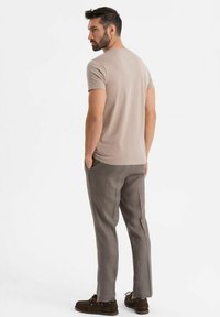 Béžové přiléhavé tričko ve spojení se šedými kalhotami, které vykazují hladkou texturu. Model stojí s rukama v kapsách a je otočen mírně dozadu.