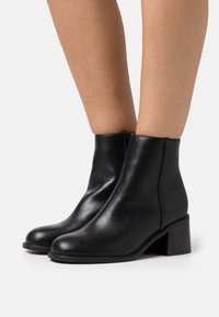 Bottines en cuir noir avec un bout arrondi et un talon bloc de hauteur moyenne. Texture lisse avec des détails de couture minimaux.