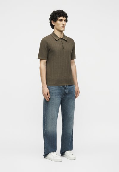 Άνδρας με σγουρά μαλλιά στέκεται, φοράει καφέ κοντομάνικο πλεκτό πόλο, μπλε τζιν με ίσια μπατζάκια και λευκά αθλητικά παπούτσια μπροστά σε απλό φόντο.