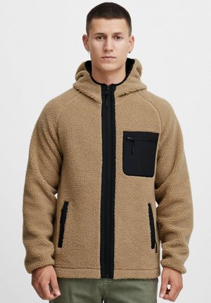 Brun fleecejakke med hette, som har en svart glidelås skulderlomme og svarte detaljer på side lommene. Myk tekstur og tettsittende passform.
