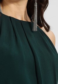 Haut dos nu vert foncé en tissu texturé, avec fronces au niveau de l'encolure et boucles d'oreilles à pampilles argentées.