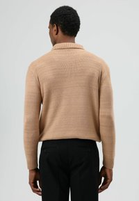 Gebreide trui in lichtbeige met een geribde kraag. Voorzien van lange mouwen en een ontspannen pasvorm, gecombineerd met zwarte, op maat gemaakte broek.