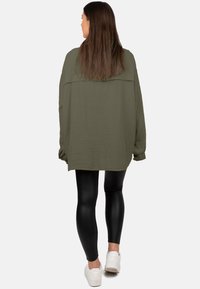 Oversized olijfgroene shirt van gestructureerde stof, lange mouwen en details op de rugnaad, gecombineerd met zwarte leggings en witte sneakers.