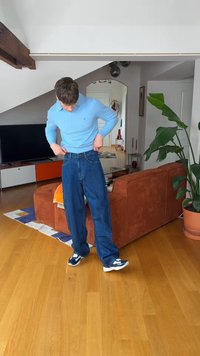 Jonge man in blauwe trui en spijkerbroek die zijn broek opricht, staande op een houten vloer in de woonkamer met een bruine bank, plant en tv.