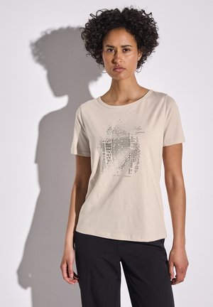T-shirt en coton beige avec un col rond, des manches courtes et un design graphique en pois noirs et texte à l'avant, associé à un pantalon noir.