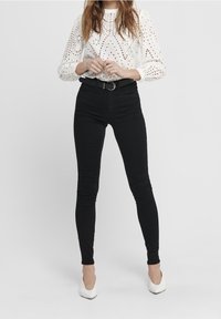 Blusa blanca de ojales con escote redondo y mangas largas. Jeans negros ajustados de tiro alto y tacones blancos de punta. Cinturón negro con hebilla redonda.