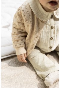 Cardigan en tricot beige avec un motif texturé, chemise blanche à boutons et pantalon vert clair avec de petits pois blancs. Tissu doux avec des poignets côtelés.