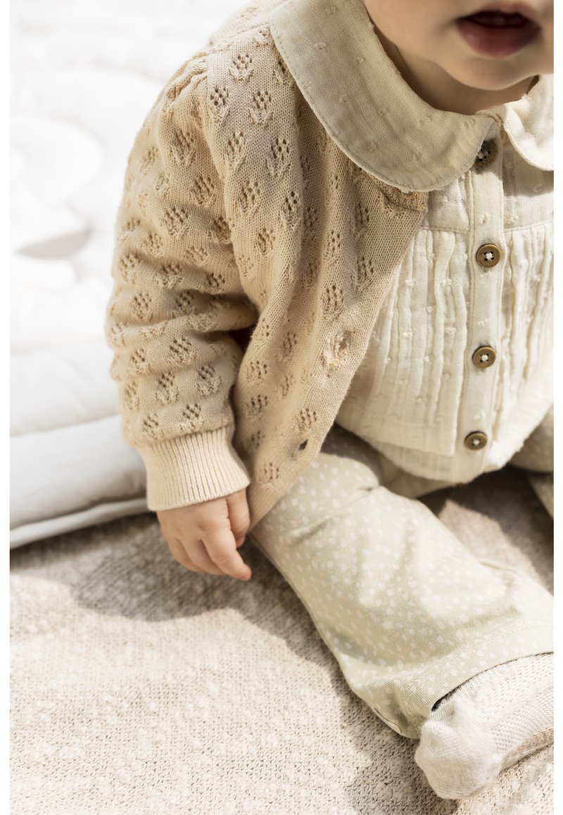 Cardigan en tricot beige avec un motif texturé, chemise blanche à boutons et pantalon vert clair avec de petits pois blancs. Tissu doux avec des poignets côtelés.