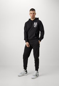 Sudadera negra con bolsillo frontal y logo gráfico, combinada con pantalones de chándal negros. El modelo lleva zapatillas deportivas blancas y grises. Diseño simple, ajuste casual.
